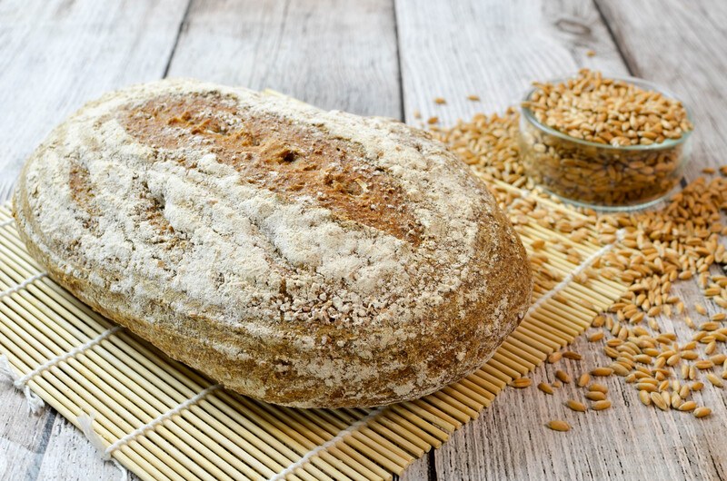 on rustic wooden surface is a spelt bread resting on top of a wooden placemat with a small dish full of spelt grain with loose spelt grain around it