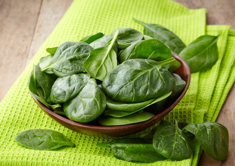 on a wooden surface is a dark colored bowl full of spinach, resting on a green kitchen towel, with loose spinach around it