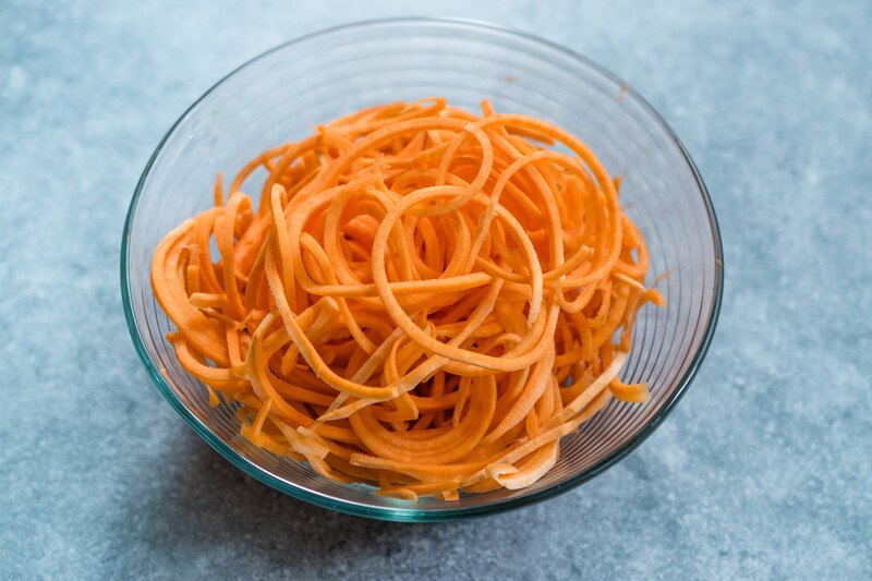 on a bluish concrete surface is a clear bowl with spiralized sweet potatoes