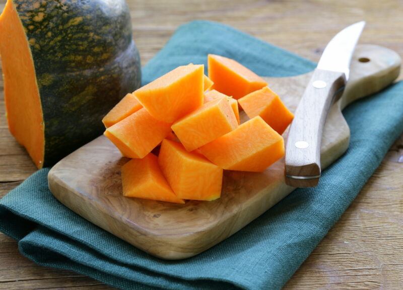 on a wooden surface is a wooden chopping board with cubed squash and knife, resting on a blue table napkin with a big chunk of squash beside it