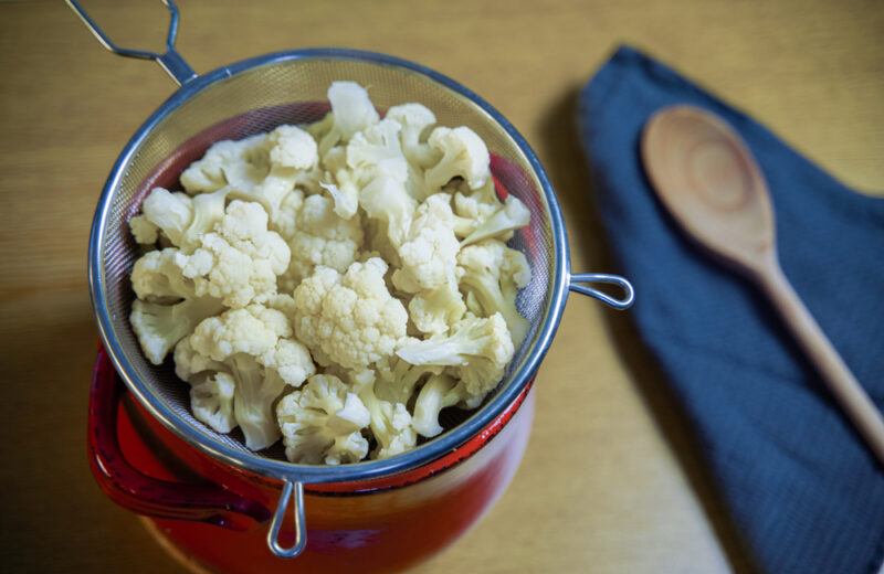 steamed cauliflower florets in a metal colander with a wooden ladle on a blue table napkin beside it