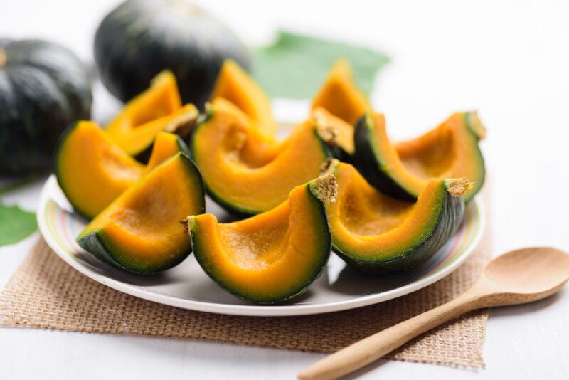 a white plate with steamed squash, resting on top of a brown place mat and wooden spoon beside it, at the back are a couple of whole squash