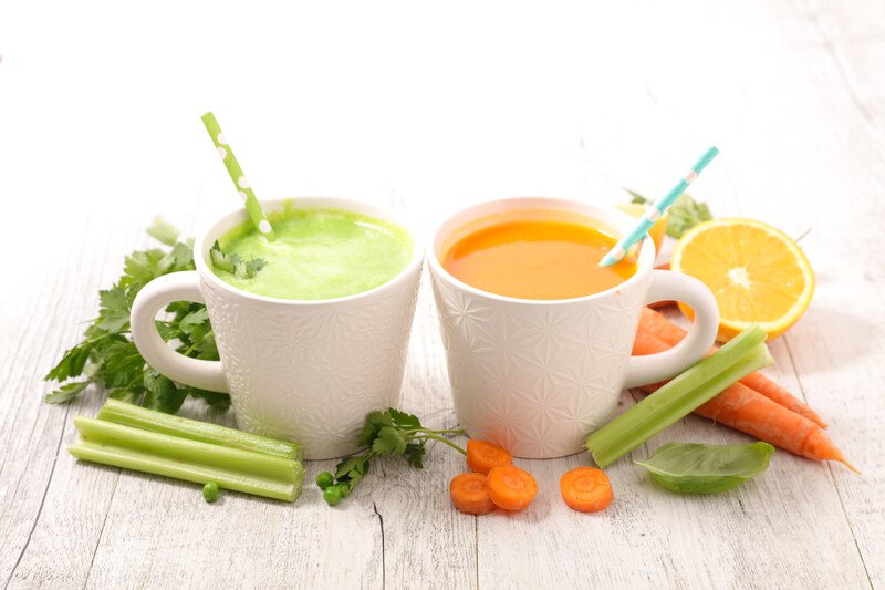 on a white wooden surface is a couple of white cup full of green and orange strained vegetable juice, with fresh vegetables around it