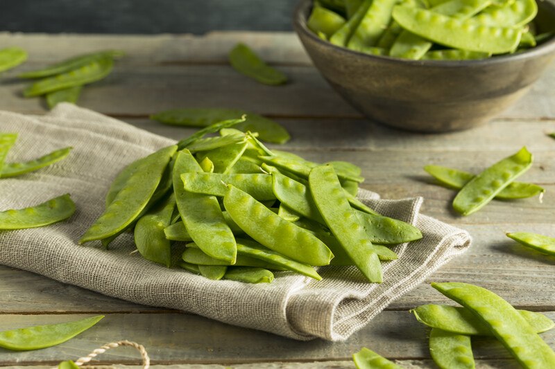 on a wooden surface is a closeup image of a pile of sugar snap peas on a tan colored table napkin, with loose sugar snap peas around it and a bowl full of sugar snap peas at the back