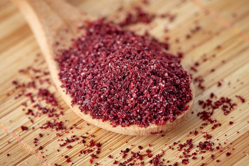 on a wooden surface is a closeup shot of a wooden ladle full of sumac, with loose sumac around it