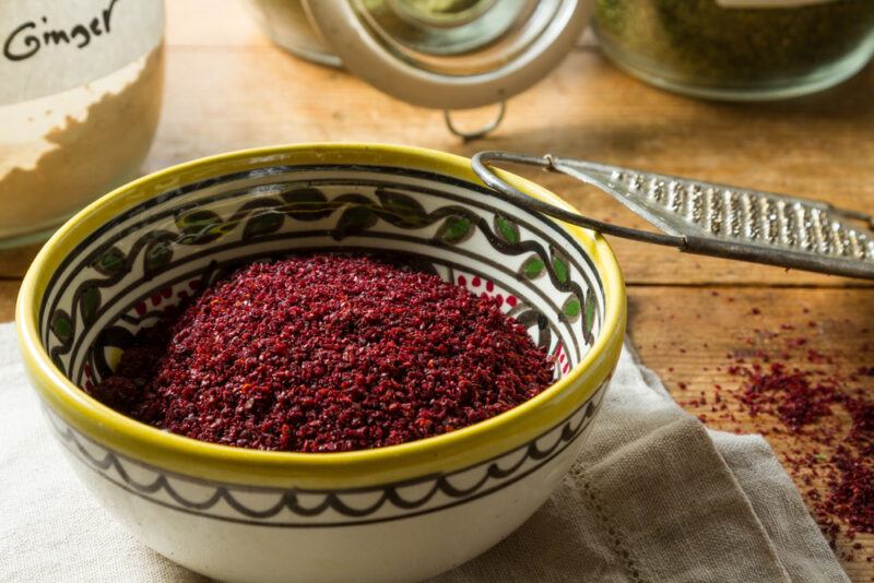 a white bowl with yellow rim and green prints full of sumac spice resting on a tan colored table napkin, beside it is a metal grater