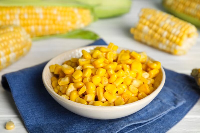 a white bowl full of corn kernel resting on a dark blue denim table napkin, around it are corn on the cobs