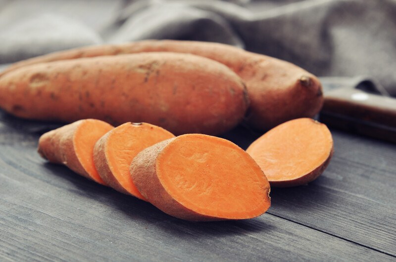 closeup image of a wooden surface with a couple of whole sweet potatoes and a few sliced ones