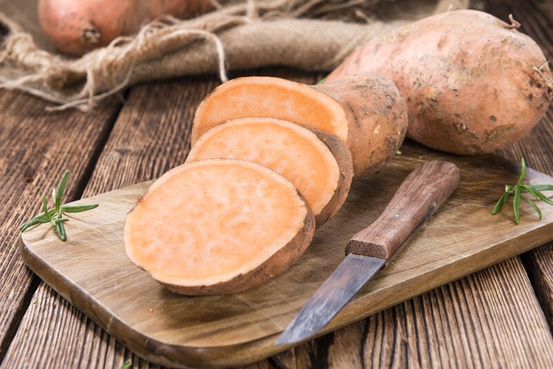 on a rustic wooden surface is a wooden chopping board with whole and sliced sweet potatoes with a knife beside it, behind it is a burlap with whole sweet potatoes