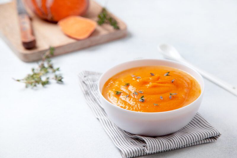 on a white concrete surface is a white bowl full of sweet potatoes puree, resting on top of a white with grey stripes table napkin, beside it is a long white ceramic spoon, at the back is a wooden chopping board with sliced sweet potatoes, knife, and herbs on it