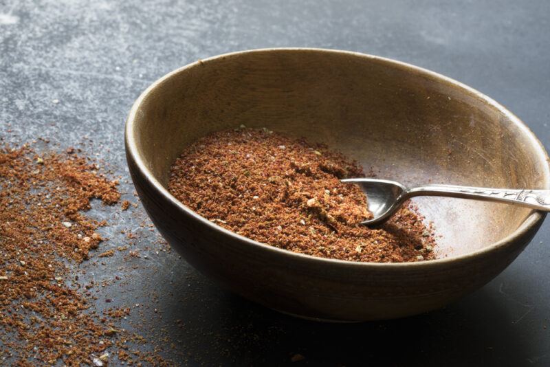 on a grey concrete surface is a wooden bowl with taco seasoning and a silver spoon, beside it are loose taco seasoning powder