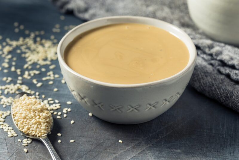 on a dark wooden surface is a white bowl of tahini with a silver spoon beside it full of sesame seeds, around are loose sesame seeds as well