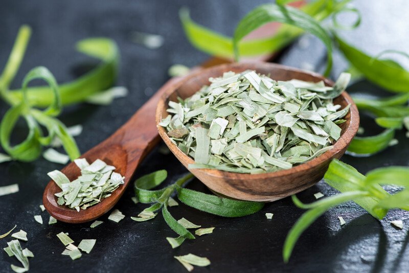 on a dark surface is a wooden bowl full of dried tarragon with wooden spoon beside it with dried tarragon and fresh tarragon leaves around it