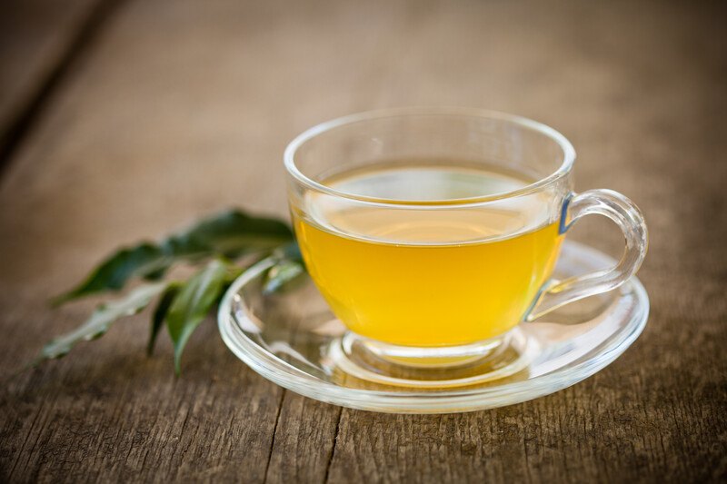 on an aged wooden surface is a clear cup and saucer with green tea, with a sprig of fresh green tea leaves beside it