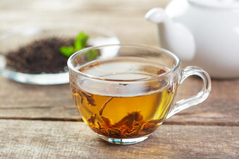 on a rustic looking wooden surface is a clear glass cup of tea, at the back is a small glass dish of dried tea leaves and a white ceramic tea pot