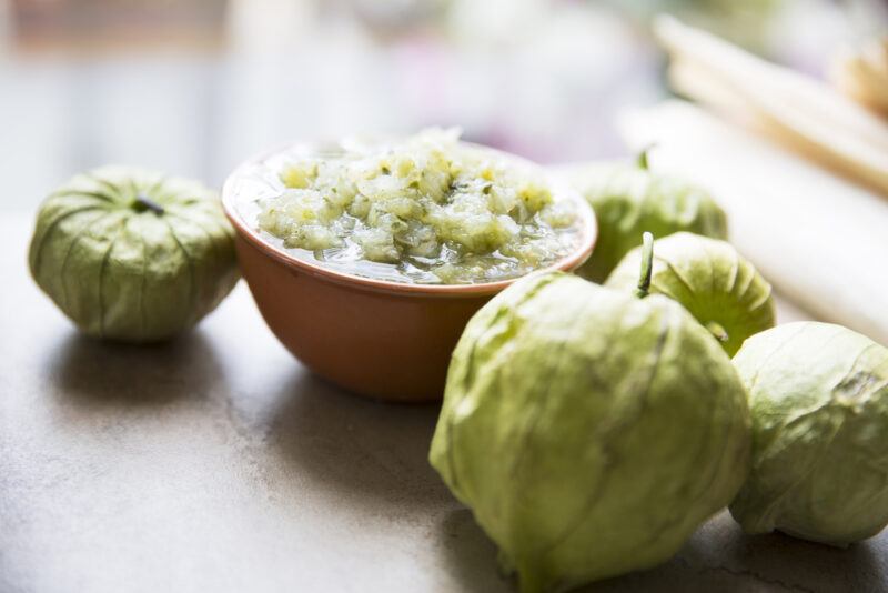 closeup image of tomatillos on a wooden surface with a small bowl of salsa verde at the back