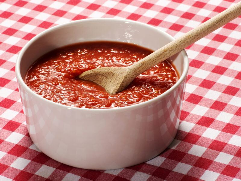 on a checkered white and red table napkin is a white bowl full of tomato puree with a wooden spoon in it