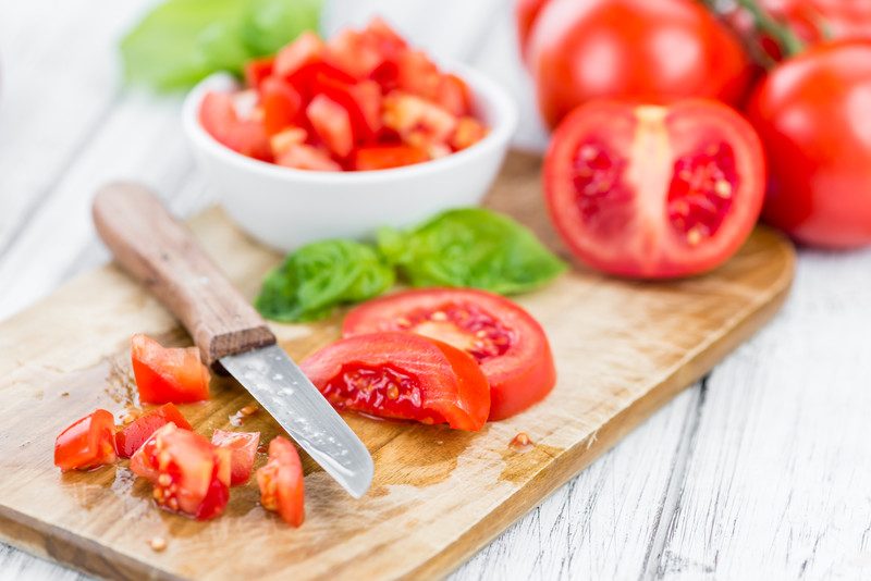 a closeup image of diced tomatoes on a wooden chopping board with tomatoes, herbs, and a bowl of diced tomatoes at the back