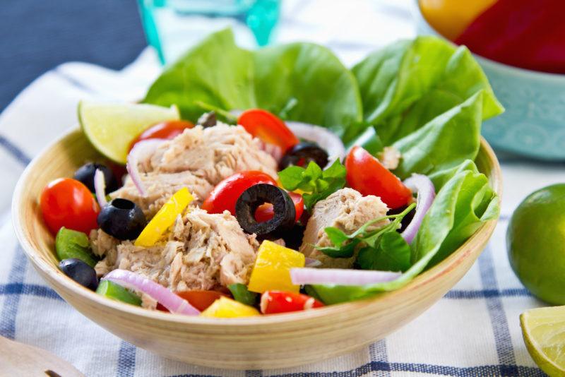 a wooden bowl filled with tuna salad containing chinks of tuna, lettuce, onions, tomatoes, and olives