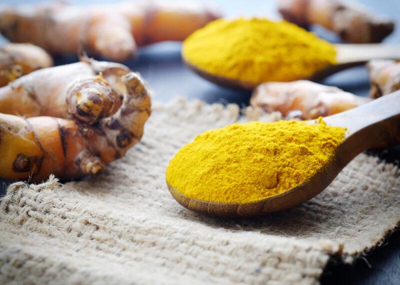 a closeup image of a wooden spoon full of turmeric powder, resting on a burlap with fresh turmeric rhizomes beside it and another wooden spoon of turmeric powder at the back