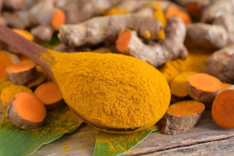 on a rustic looking wooden surface is a spread of fresh turmeric rhizomes with a couple of leaves and a wooden spoon full of turmeric powder