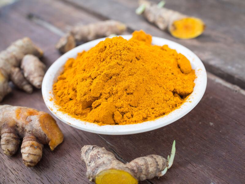 on an aged looking wooden surface is a white bowl full of turmeric powder with loose fresh turmeric rhizomes around it