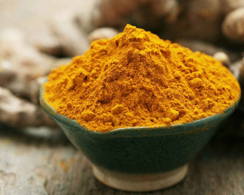 on a wooden surface is a closeup image of a green bowl with a heaping mound of turmeric powder with fresh turmeric rhizomes  at the back