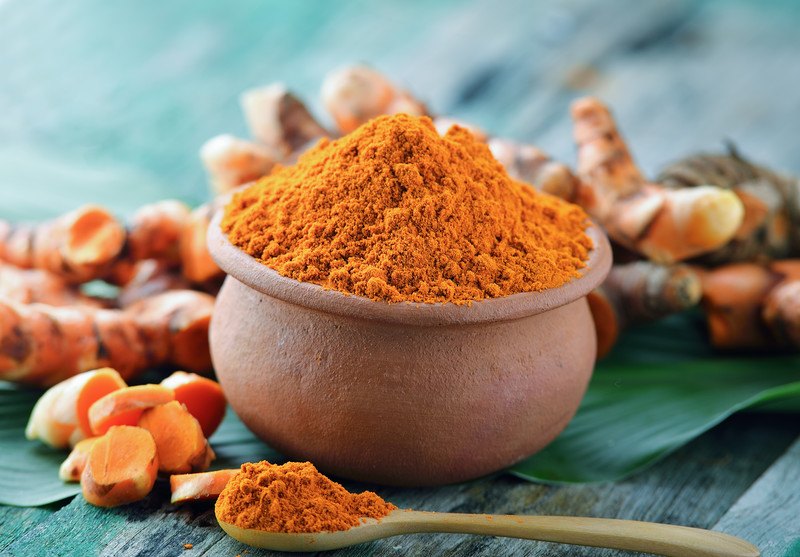 a small clay pot full of turmeric resting on a banana leaf with fresh turmeric around it and a wooden spoon full of turmeric powder in front of it