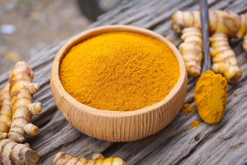 on an aged wooden surface is a light brown clay bowl full of turmeric powder, with fresh turmeric rhizomes and wooden spoon full of turmeric powder beside it