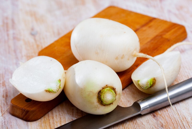 on a light brown wooden surface is a wooden chopping board with fresh turnips on it and a knife beside it