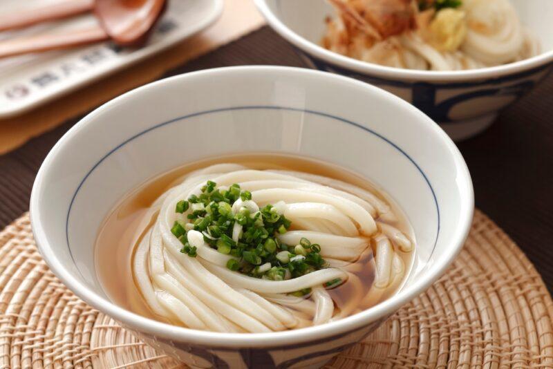 a closeup image of white bowl with blue lines around the rim containing udon noodle soup with chopped greens, resting on a place mat made from jute