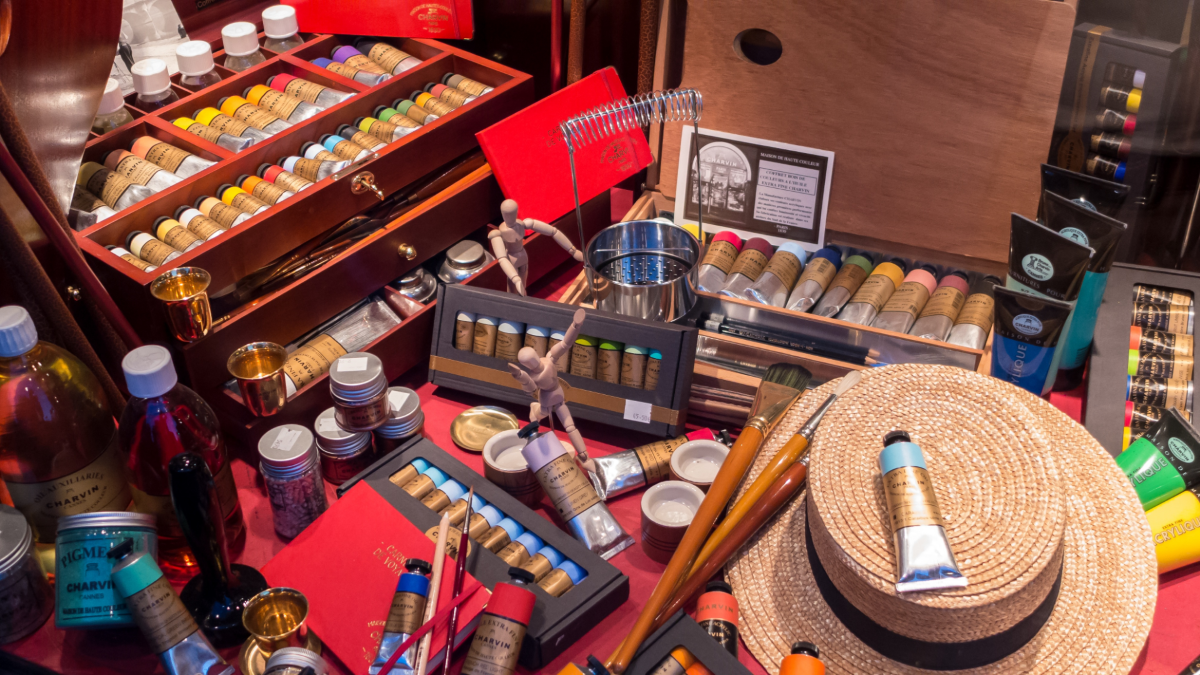 Painting supplies covering a  table with a straw hat in the lower right corner with a tube of paint on top and brushes sitting at brim