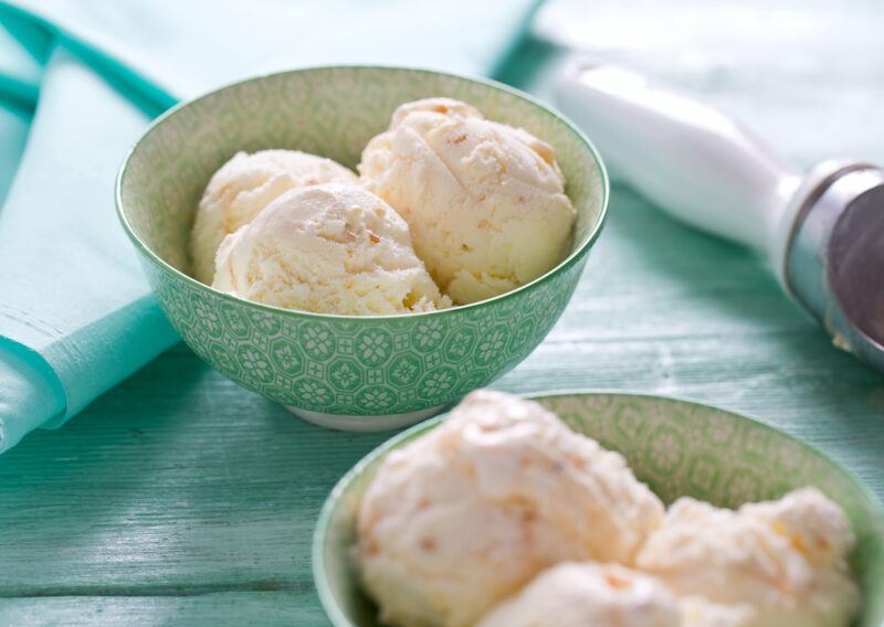 on a greenish wooden surface is a couple of bowls with green prints, both bowls with vanilla ice cream, beside one of the bowl is an ice cream scooper and a green table napkin