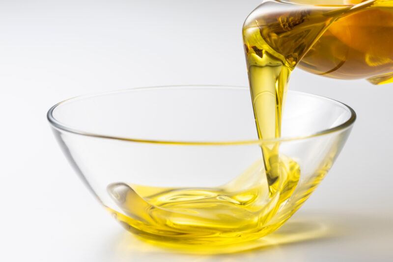 closeup image of a clear glass bowl with vegetable oil getting poured in it from a container that can be partially seen at the back