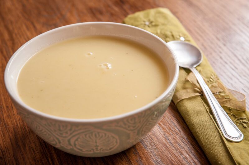 on a wooden surface is a white bowl with veloute, beside it is a silver spoon resting on top of a yellow table napkin