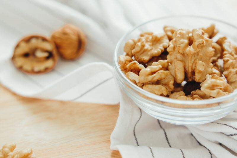 on a wooden surface is a clear glass bowl of walnuts resting on top of a white table napkin with grey stripes, around are loose walnuts