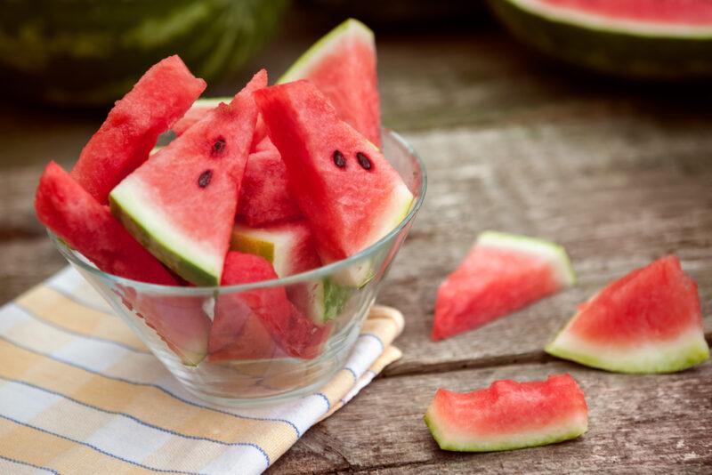 on a rustic looking wooden surface is a clear glass bowl of sliced watermelon resting on top of a striped white, yellow, and grey table napkin, loose watermelon slices beside it