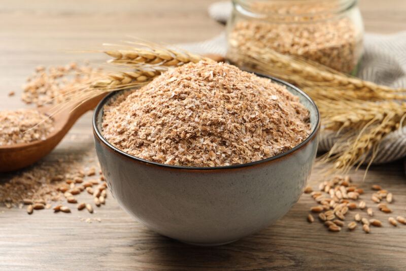 on a wooden surface is a bowl full of wheat bran with loose wheat bran, wooden ladle and wheat behind it