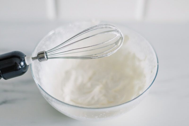 a closeup image of a metal whisk, on a white surface is a glass bowl with whipped cream