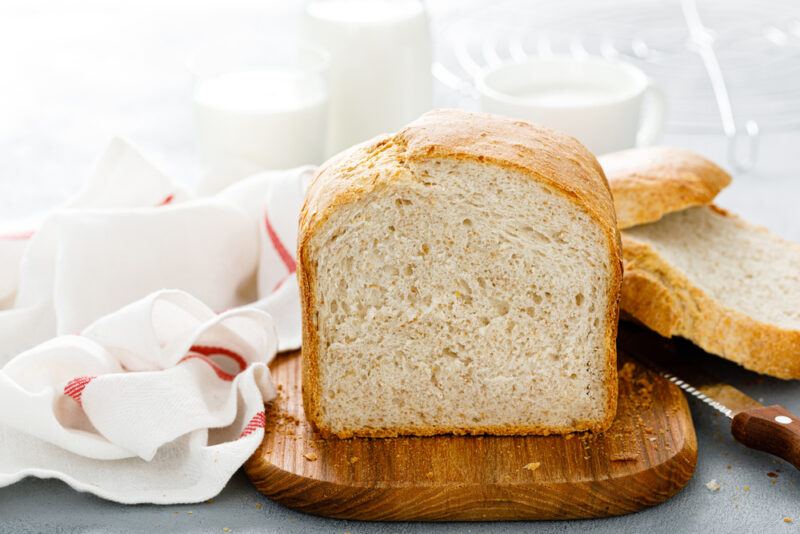 on a wooden board is a sliced loaf of white bread with a knife beside it and a white table napkin with red stripes at the back along with a couple glass and one cup of milk