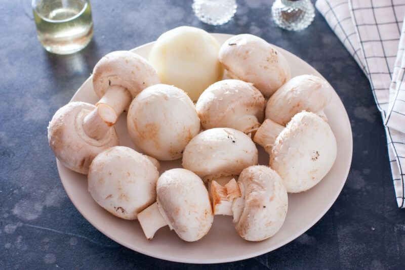 on a dark concrete surface is a white plate full of white button mushrooms with a table napkin beside it and a small jar of oil and salt and pepper shaker at the back