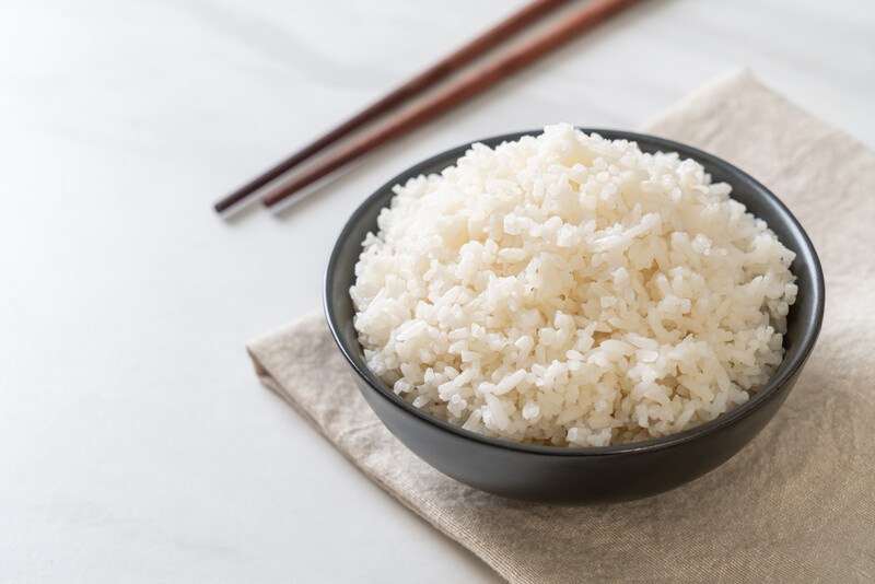 on a white surface is a black bowl full of cooked white rice, resting on a light brown table napkin with chopsticks at the back