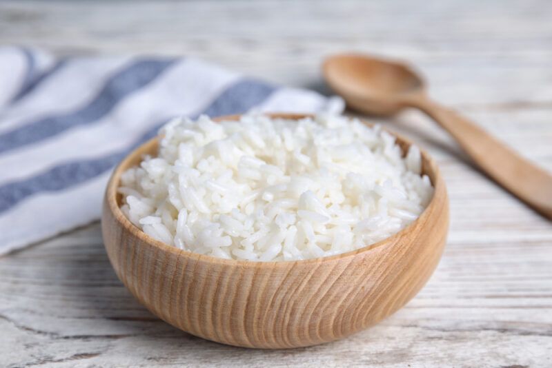 on a light colored aged wooden surface is a wooden bowl of white rice with a wooden spoon and white and blue table napkin behind it