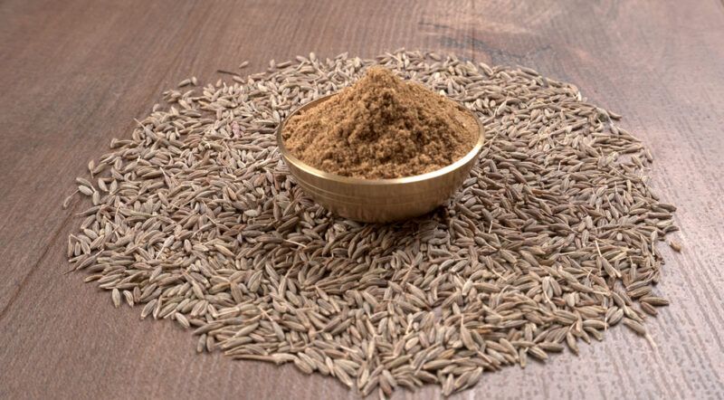 on a wooden surface is a small mound of whole cumin seeds with a small gold bowl on top of it full of ground cumin seeds