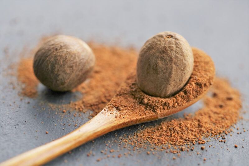 on a dark surface is a wooden ladle with whole and ground nutmeg with loose nutmeg powder and one whole nutmeg around it