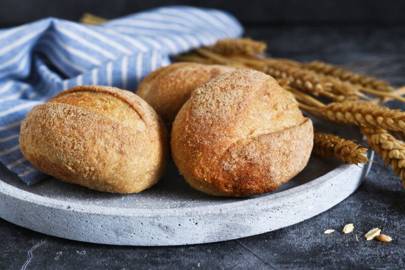 on dark concrete surface is a closeup image of freshly baked whole grain bread with oat stalks beside it, resting on a round stone dish with a white and blue striped table napkin behind it