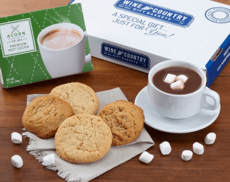 Wine and Country hot cocoa gift set with acorn bakery cookie box with four cookies sitting on a napkin and a white mug and saucer with hot cocoa in it, plus mini marshmallows in the cup and scattered on the table