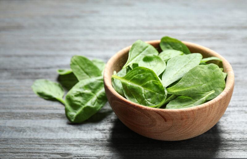 on a black wooden surface is a light brown wooden bowl full of spinach, with loose spinach beside it