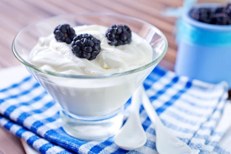 on a wooden surface is a clear bowl of yogurt with three blackberries on top, resting on striped white and blue table napkin, at the back is a blue container with blackberries