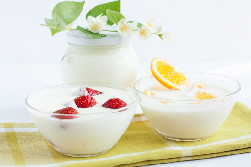 on a yellow table napkin are a couple of clear bowls with yogurt, one topped with strawberries, the other one with sliced oranges, with a jar full of plain yogurt with a stem with leaves and tiny white and yellow flowers at the back